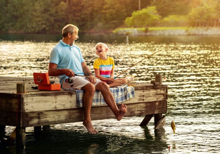 A man and a child sit on a dock with fishing poles laughing