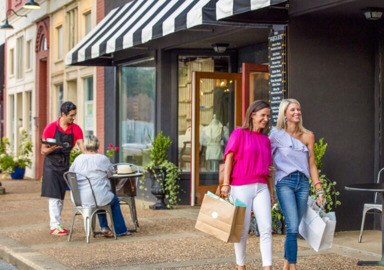 Two women walk with shopping bags, while a waiter takes an order at a cafe in the background