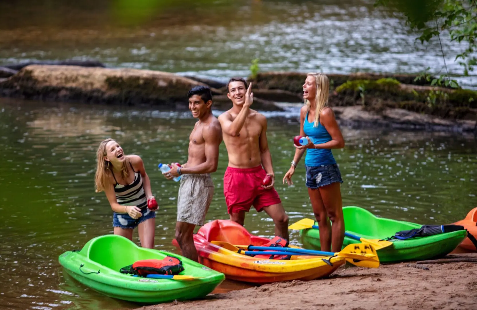 A group of four young, fit people stand talking and laughing beside their kayaks