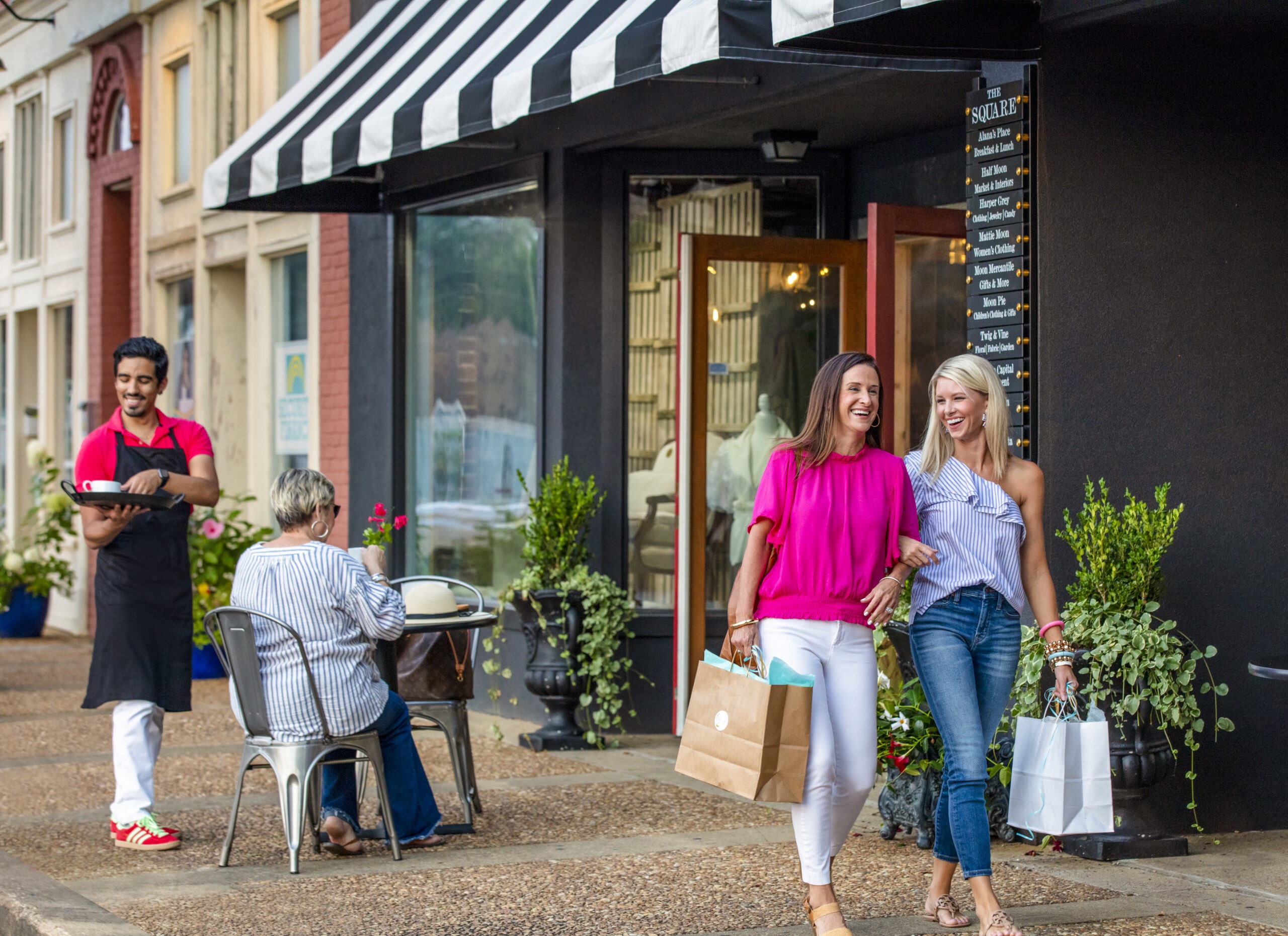 Two women with shopping bags stroll a downtown street, with a cafe patio seen behind them