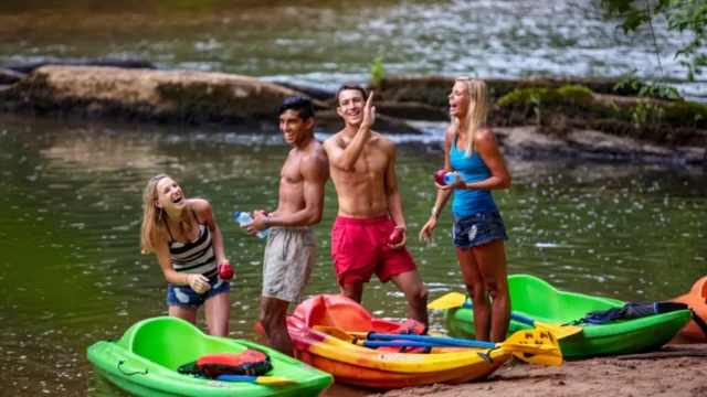 A group of four young, fit people stand talking and laughing beside their kayaks