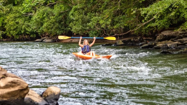 A woman in a kayak holds her paddle over her head triumphantly